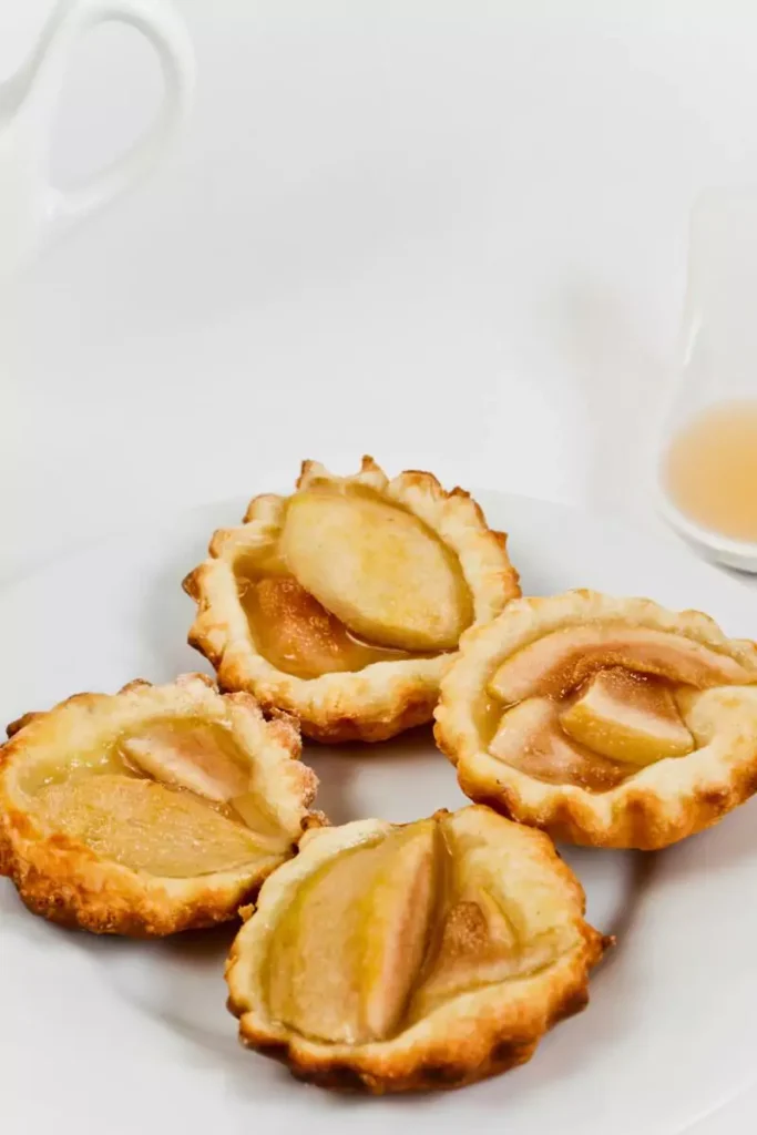 Golden-brown mini phyllo tartlets filled with creamy blue cheese and diced pear, garnished with fresh herbs on a white serving plate.