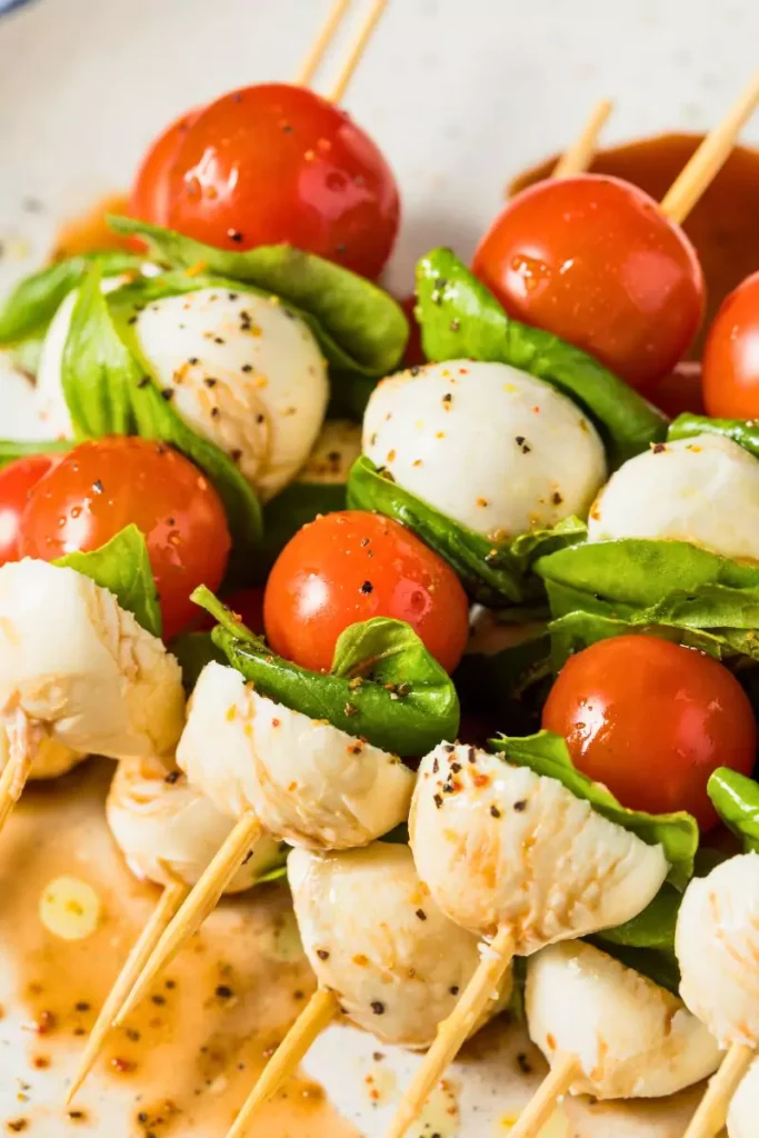 Close-up photo of Caprese skewers with grape tomatoes, mozzarella cubes, and fresh basil on toothpicks, arranged on a white platter