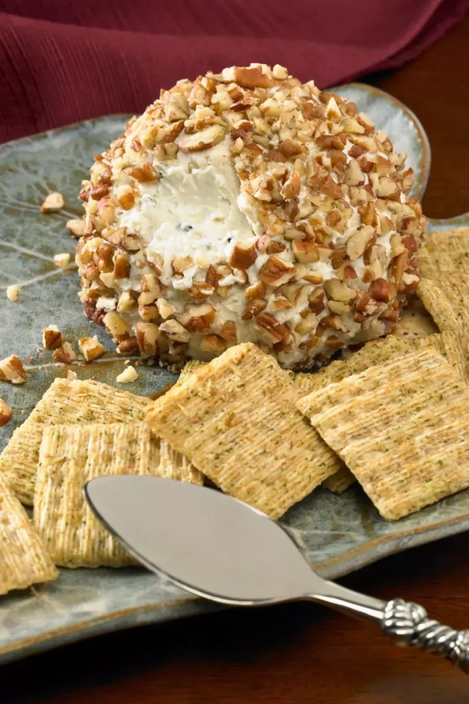 A pine cone-shaped cheese ball garnished with fresh rosemary and covered with almonds, displayed on a white plate.