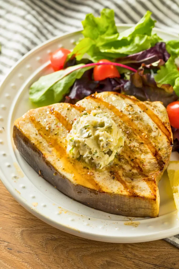 A beautifully plated Swordfish a la Siciliana with raisins, olives, capers, and pine nuts, served alongside a vibrant green salad.