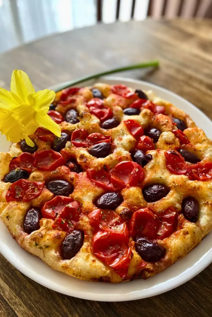 A golden-brown round focaccia bread topped with olives, sun-dried tomatoes, and herbs, fresh out of the oven on a wire rack.