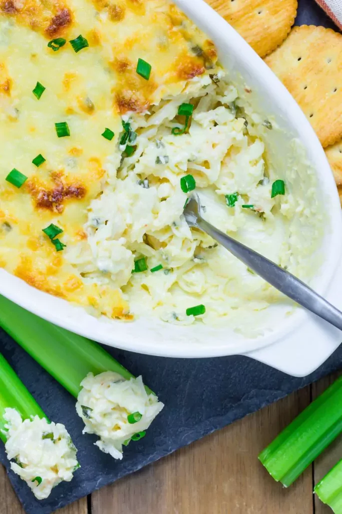 A warm, cheesy baked crab dip served in a tray, garnished with sliced green onions and surrounded by fresh vegetables and crackers.