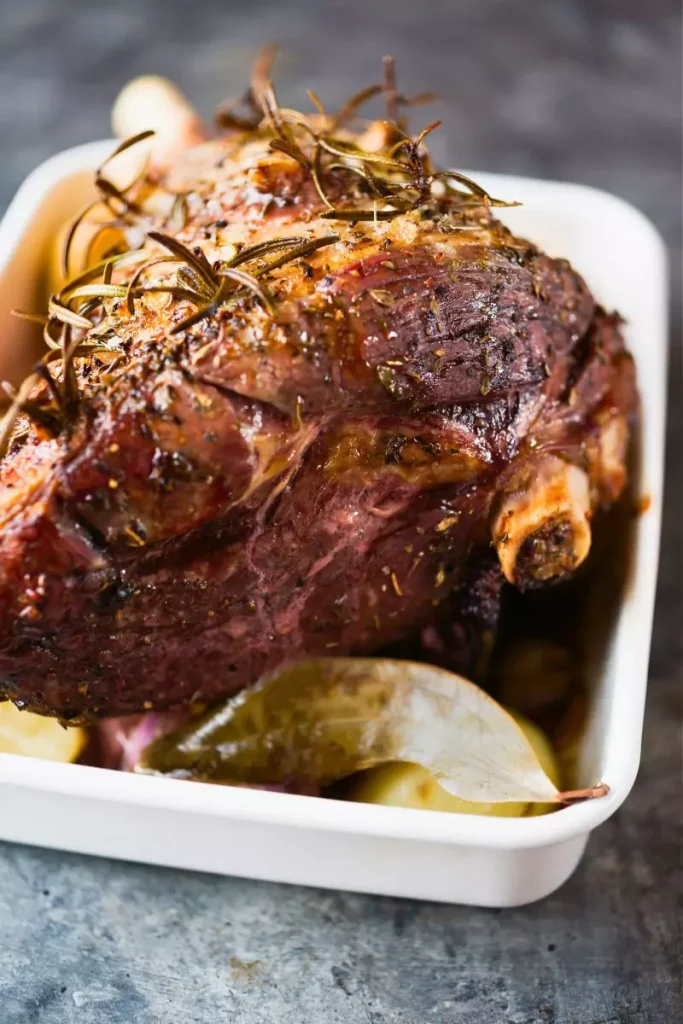 A beautifully roasted leg of lamb garnished with fresh herbs, surrounded by artichokes and lemon halves in a rustic serving dish.