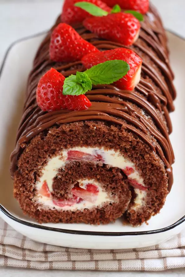 A roll of chocolate Swiss roll with creamy filling, dusted with strawberries, served on a white plate.