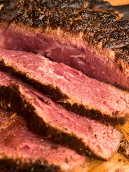 Sliced prime rib with a golden crust served on a wooden board, surrounded by roasted vegetables and a bowl of au jus.