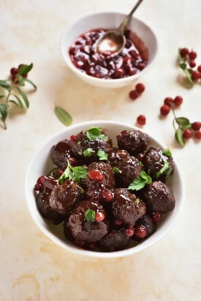 Close-up of cranberry sauce meatballs in a tangy red sauce, garnished with fresh herbs, served in a white bowl for a holiday appetizer.