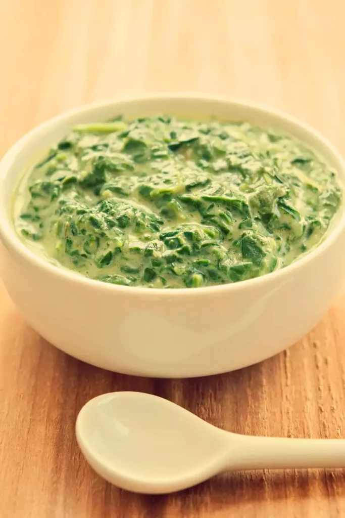 A bowl of creamed spinach served in a white dish, garnished with freshly grated parmesan cheese and a sprinkle of ground pepper.