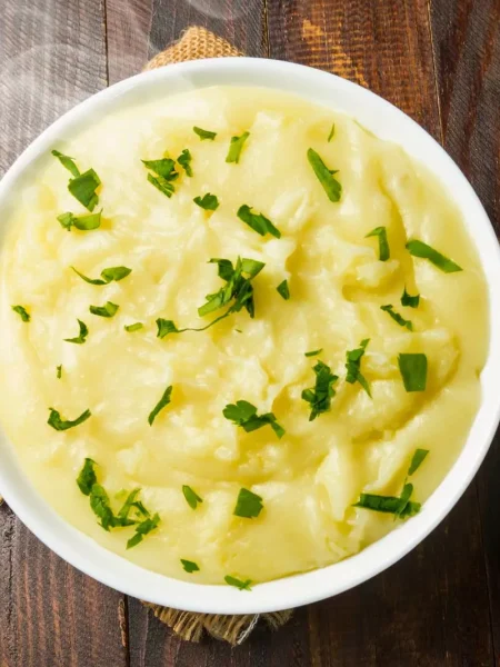 A bowl of creamy mashed potatoes topped with melted butter, garnished with chopped parsley, served on a rustic wooden table.