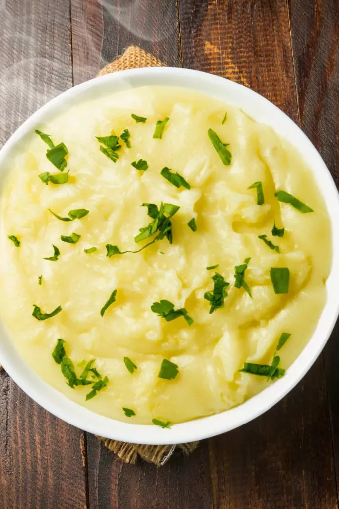 A bowl of creamy mashed potatoes topped with melted butter, garnished with chopped parsley, served on a rustic wooden table.