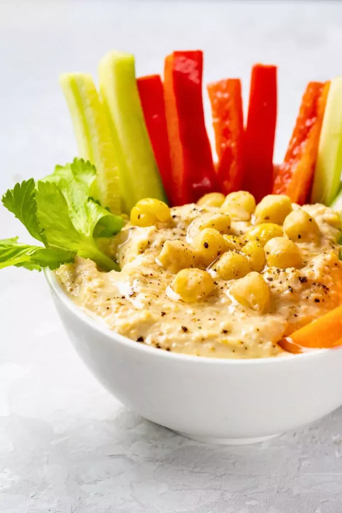 A bowl of creamy hummus garnished with olive oil and spices, surrounded by pita bread and fresh vegetables.