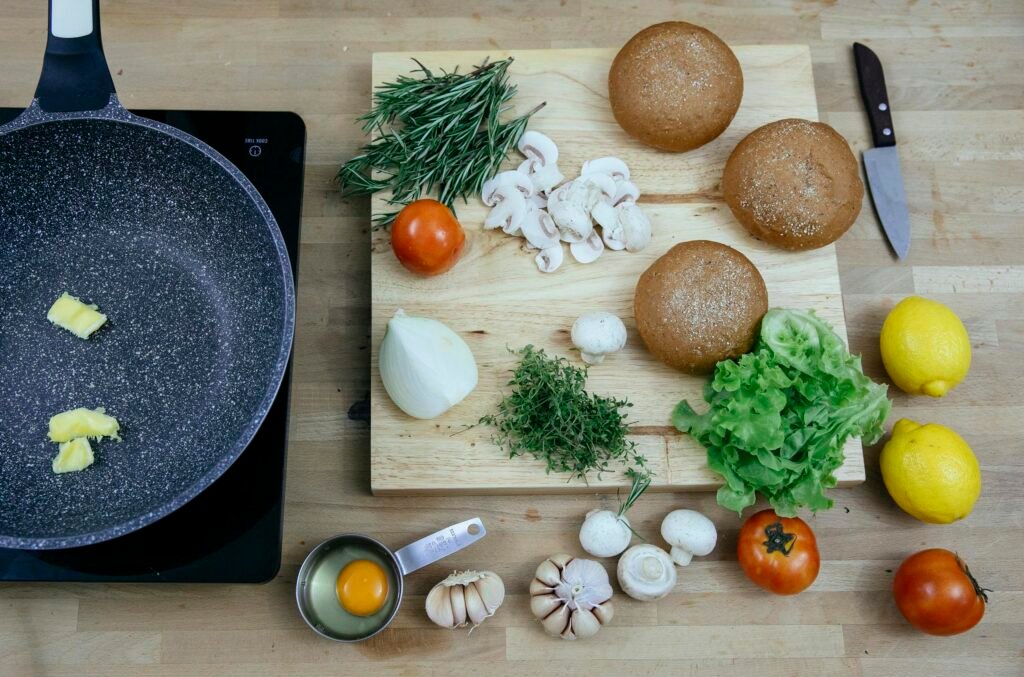 Top view of pan with butter on stove placed near wooden board with buns greens and fresh vegetables in kitchen